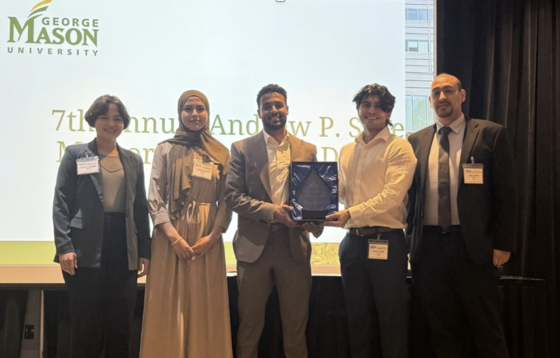Five people stand in front of a conference room, with two in the middle holding a prize for first place